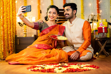 Happy indian couple taking selfie on mobile phone by holding flowers plate during diwali festival celebration at home - concept of togetherness, social media sharing and traditional culture. - obrazy, fototapety, plakaty