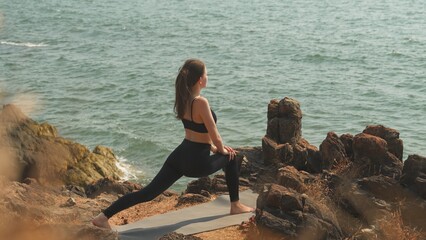 young woman is doing Ashva Sanchalanasana on a seaside rocky shore