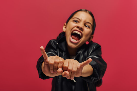Blurred and mad preadolescent girl in stylish leather jacket showing rock gesture at camera while standing and posing isolated on red, girl with cool and contemporary look