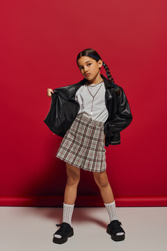Full length of fashionable preadolescent girl with hairstyle posing in leather jacket and checkered skirt and looking at camera while standing on red background, stylish preteen outfit concept