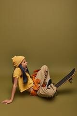 Side view of excited preadolescent girl with colorful hair wearing trendy urban outfit and hat while sitting near skateboard on khaki background, girl with cool street style look