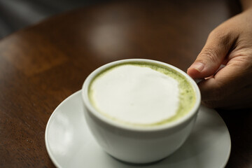 Closeup hand holding white cup of hot green tea latte on wood background in restaurant.Best of menu in the coffee shop.Soft focus.