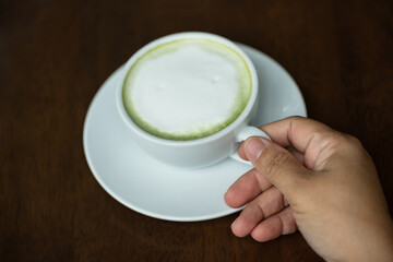 Closeup hand holding white cup of hot green tea latte on wood background in restaurant.Best of menu in the coffee shop.Soft focus.