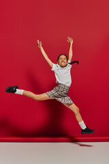Fototapeta na wymiar Excited and cheerful preteen girl in white t-shirt and stylish checkered skirt jumping and having fun while posing on red background, hairstyle and trendy accessories concept