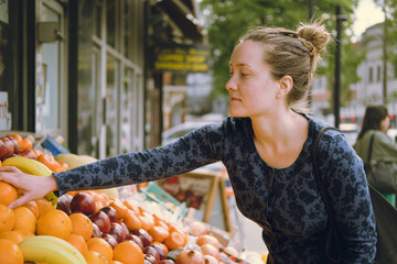 Side view of young-adult woman taking fruit with her hand when shopping at a food market, choosing...