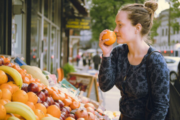 Side view of young-adult woman taking fruit with her hand, smelling an orange when shopping at a food market, choosing what to buy at the street fruit stand, carrying a black tote bag. Lifestyle.