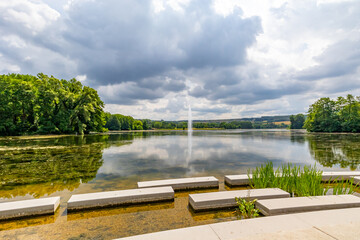 Fototapeta premium Concrete platforms on shore of Lake Echternach with waterjet gushing from a fountain, green leafy trees in background, reflection on water surface, panoramic landscape, summer cloudy day in Luxembourg