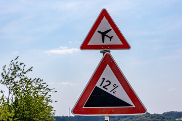 Traffic signs indicating: airport warning sign or low flying airplane and  steep 12 percent downhill gradient in road, against blue sky on a sunny summer day in Germany