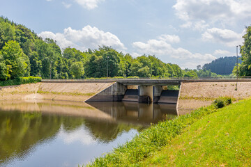Fototapeta na wymiar Staumauer Bitburger dam on river Prum, reflection on water surface, vehicular bridge, green leafy trees in background, Stausee Bitburg reservoir on a sunny spring day in Germany