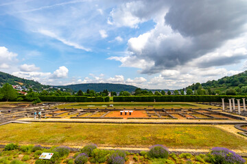 Fototapeta na wymiar Panoramic landscape of restored ruins of Roman villa in Echternach lake park, small stone walls, pillars, green trees, village and mountains in the background, sunny summer day in Luxembourg