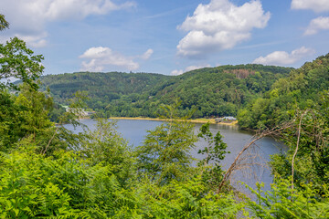 Fototapeta na wymiar Partial view of Stausee Bitburg reservoir lake through green foliage, calm water, mountains with abundant trees with green foliage in background, sunny spring day with blue sky in Germany