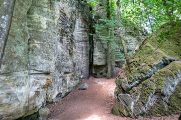 Huge rock formations in nature reserve Teufelsschlucht, walls with moss, uneven texture and grooves, hiking trail, trees with green foliage in background, sunny summer day in Ernzen, Germany