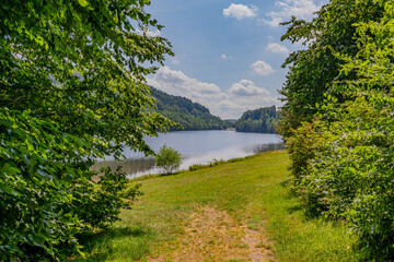 Path descending between wild vegetation towards  Stausee Bitburg lake and mountains with abundant trees with green foliage in background, sunny spring day with a blue sky in Germany