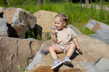 Sitting on large granite stones in the park, a girl eats strawberries on a hot summer day. The girl eats berries rich in vitamins. The concept of a child having a snack while walking in the park.