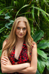 Glamorous blonde model in stylish black swimsuit and transparent robe posing in lush green tropical greenhouse
