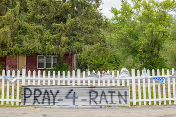 Pray 4 Rain painted on metal siding along a picket fence with a red building and trees in the background.