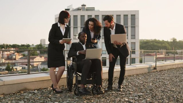 African american executive in wheelchair checking business plan on remote laptop together with group of foreign investors outdoors. Diverse colleagues with devices bending to provide additional info.