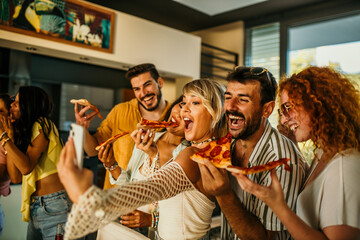 Friends eating a pizza, taking a group selfie and having fun during a house party.