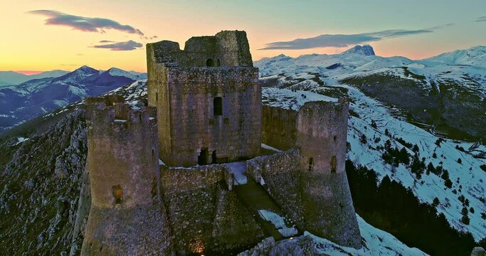 Aerial view of the Rocca di Calascio with snow and illuminated by the light of the sunset, behind the chain of the Gran Sasso and the Corno grande. Gran Sasso and Monti della Laga National Park, Abruz