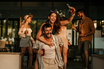 A group of young people exchange enthusiastic dancing, waving, and hugging each other, with cocktail bottles in hand going to watch a sunset.