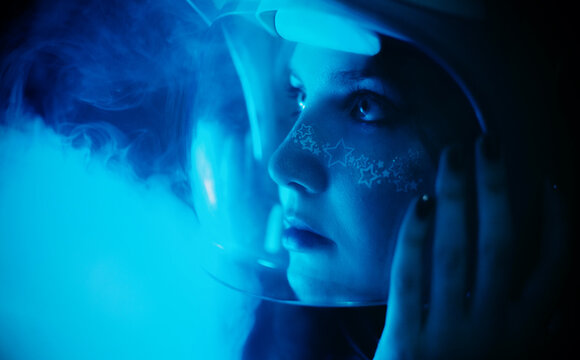 Amidst the dark of night and mist, a young woman in an astronaut helmet, adorned with starry makeup illuminated by the blue light. Exploration, cosmic space, and the limitless of human curiosity. 