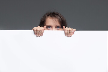 A young woman hides behind a white banner, looks out, only her eyes are visible.