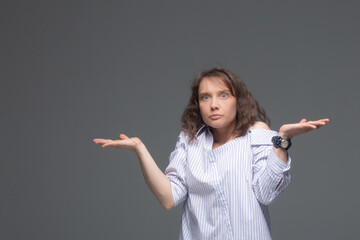 A young woman on a gray background with bewilderment on her face spreads her hands and shrugs her shoulders