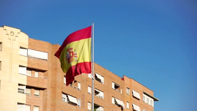 Bandera de Espa&ntilde;a ondeando al viento con un cielo azul y un bloque de pisos al fondo.