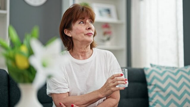 Middle age woman drinking glass of water sitting on sofa home