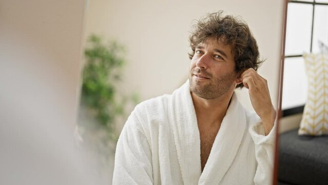 Young hispanic man wearing bathrobe cleaning ear with cotton swab looking on mirror at home