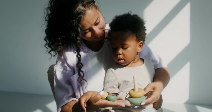 Mom with a child of African-American appearance, dressed in white clothes, playing pyramid while sitting in a chair in a white room