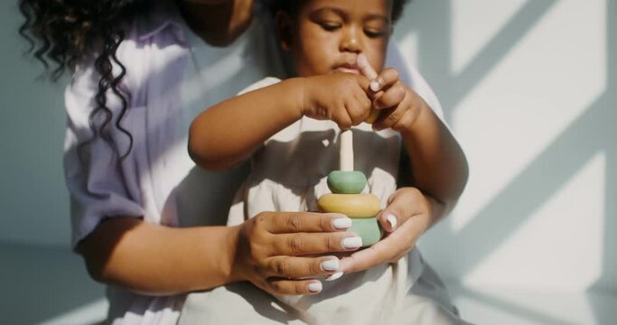 Mom with a child of African-American appearance, dressed in white clothes, playing pyramid while sitting in a chair in a white room