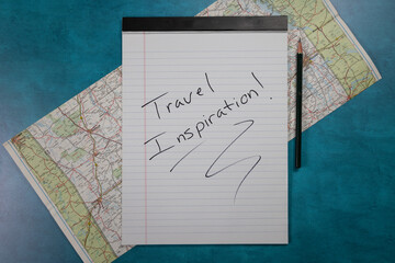 Notepad with travel inspiration hand written on it old road maps in the background lay flat