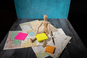 Travel themed background wooden manikin doll sitting on old road maps holding pencil with blank posted notes