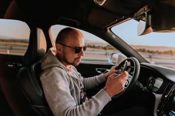 A man with a sunglasses driving a car and type a message on smartphone at sunset. The concept of car travel