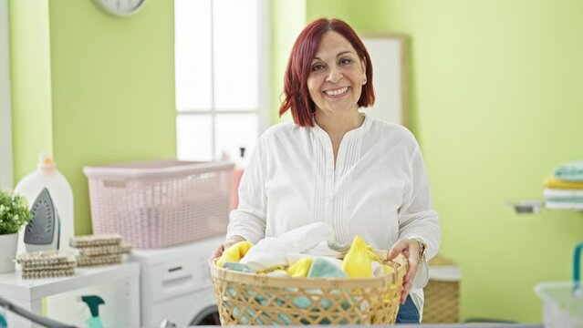 Middle age woman smiling confident holding basket with clothes at laundry room