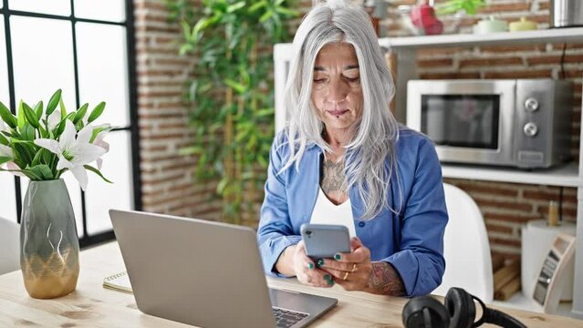 Middle age grey-haired woman using laptop and smartphone sitting on table at dinning room