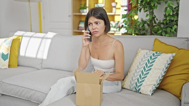 Young beautiful hispanic woman speaking on the phone unpacking cardboard box at home