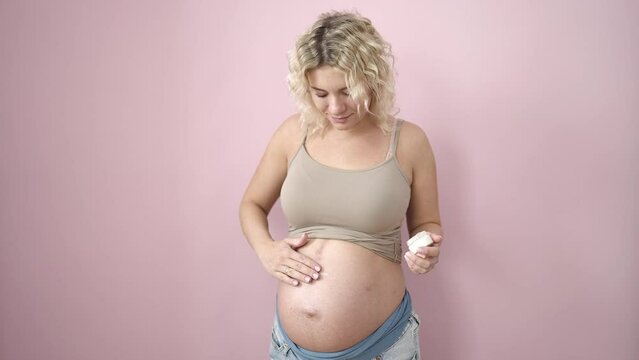 Young pregnant woman smiling confident applying skin care lotion on belly over isolated pink background