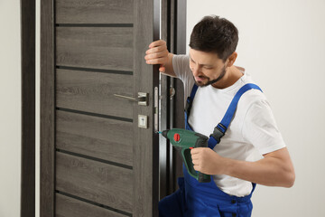 Worker in uniform with screw gun repairing door lock indoors - obrazy, fototapety, plakaty