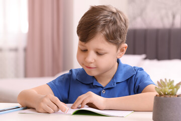 Little boy erasing mistake in his notebook at white desk indoors