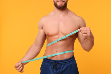 Athletic man measuring waist with tape on orange background, closeup. Weight loss concept