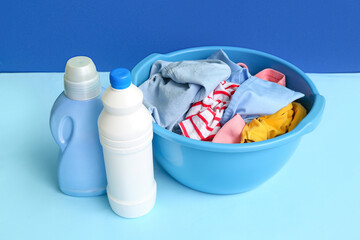 Fototapeta na wymiar Plastic basin with dirty clothes and bottles of laundry detergent on blue background