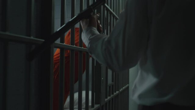 Prison guard with police baton watches criminals in prison cells. Prisoners, inmates in orange uniforms serve imprisonment terms for crimes in jail. Work in detention center or correctional facility.