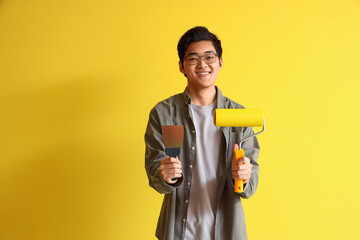 Young Asian man with putty knife and paint roller on yellow background