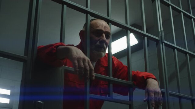 Elderly prisoner in orange uniform holds hands on metal bars in jail. Guilty criminal serves imprisonment term for crime in prison cell. Depressed inmate in detention center or correctional facility.
