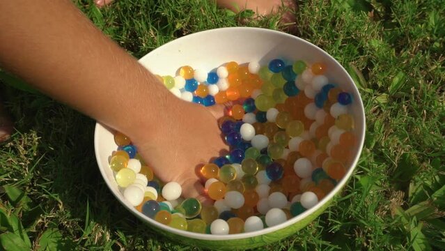 Close-up of a boy child hand touching multicolored hydrogel balls orbeez in a bowl on the grass.