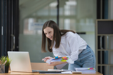 Businesswoman holding a pile of paperwork and using a laptop at her desk in an office.