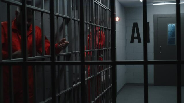 Prisoners in orange uniforms hold metal bars in jail. Guilty criminals serve imprisonment terms in prison cells. Angry inmates in detention center or correctional facility. View from jail cell.