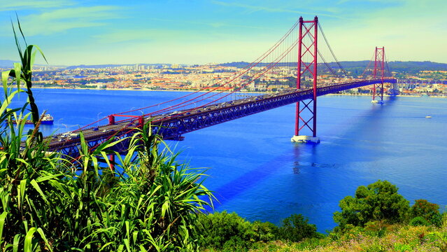  lange rote H&auml;ngebr&uuml;cke &uuml;ber den blauen Fluss Tejo (Br&uuml;cke des 25. April) in Lissabon mit gr&uuml;nem Busch 